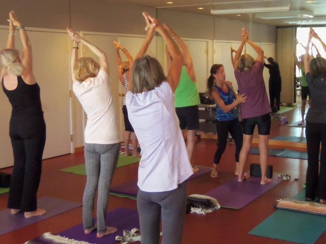 a group of people stretching in an exercise class shown from the back of the room with the instructor guiding one of the students